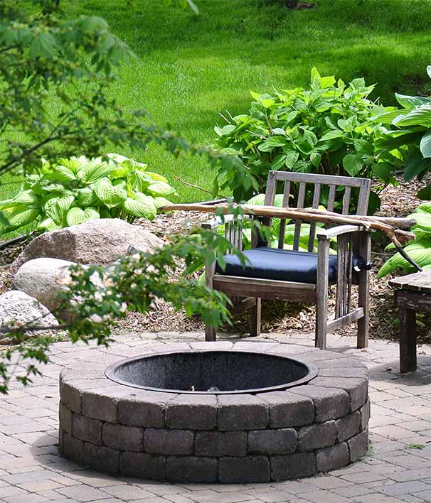A paved outdoor patio with a stone firepit and wood-framed chair with a blue seat cushion.