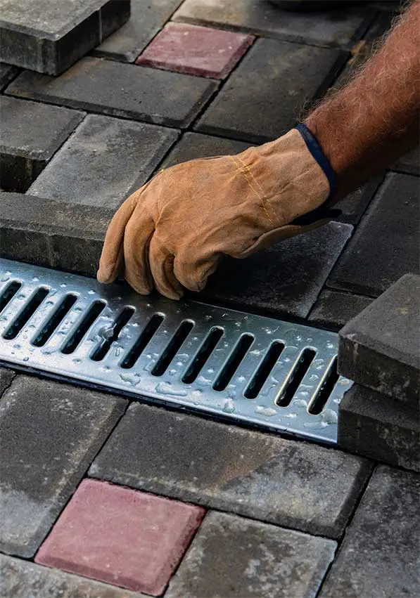 A man's hand, wearing a leather work glove, places a patio paver next to a drainage piece.