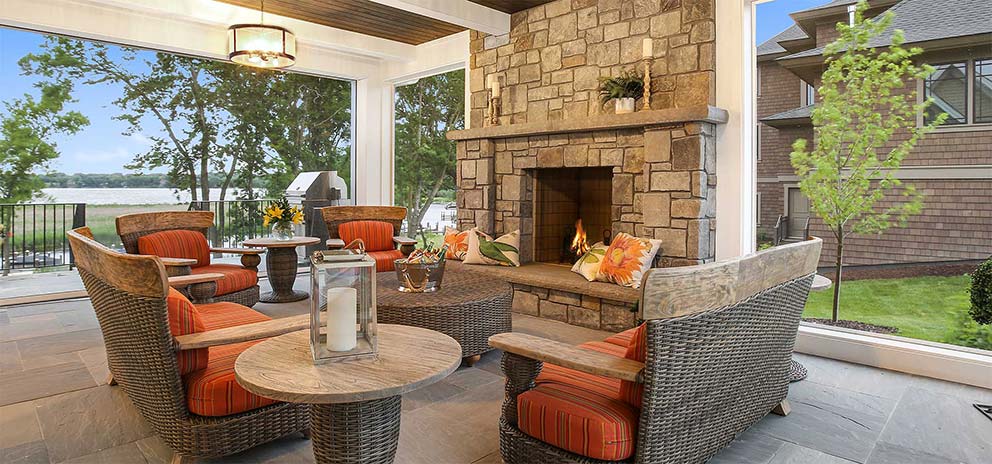 An outdoor room with comfortable seating, large stone fireplace and hanging lights overlooks a coastal waterway.